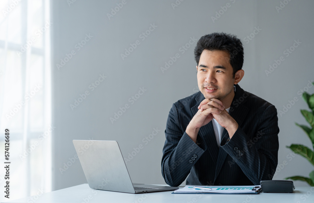 Asian young businessman sitting with his hands under his chin smiling happily after getting financial data. Statistics graphs. Income charts. while working online on laptop computer at office. Asian young businessman sitting with his hands under his chin smiling happily after getting financial data. Statistics graphs. Income charts. while working online on laptop computer at office.