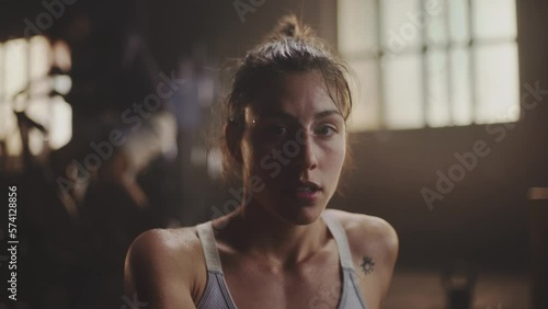 Slow-motion portrait of young beautiful sweaty woman laughing on camera in fitness centre. Close-up shot. People exercising in the background