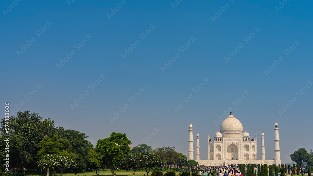Obraz premium Beautiful white marble Taj Mahal against the blue sky. Green and flowering trees grow in the garden. A lot of people are walking along the paths to the mausoleum. Copy space. India. Agra
