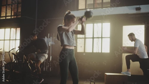 Slow-motion shot of a group of friends exercising together at gym. Woman lifting kettelbell, a man riding stationary bike, one lifting barbell and another doing jumping exercise photo