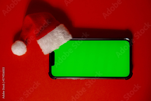 Mobile phone dressed in Santa-Claus red-white hat with chroma key screen against red background. Concept for Christmas or New Years holidays. Blank cell phone. Digital gadget wireless wishlist concept
