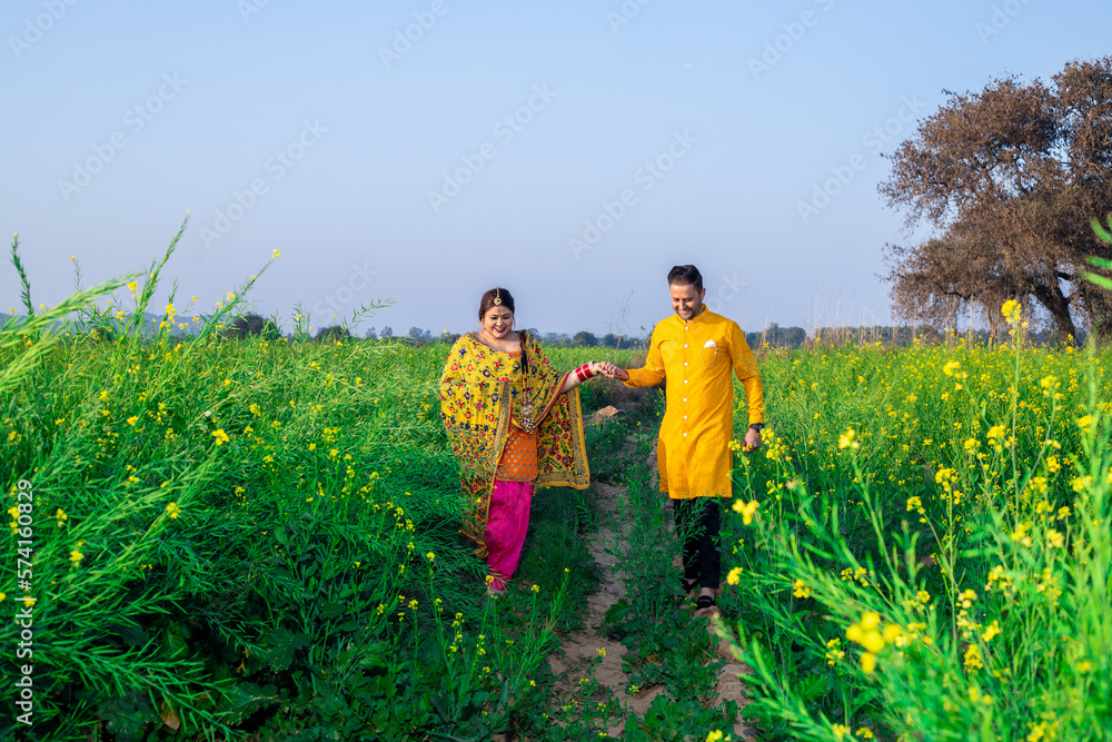 Fototapeta premium Portrait of Happy young indian punjabi couple wearing colorful outfit holding each others hand walking together in agriculture field.