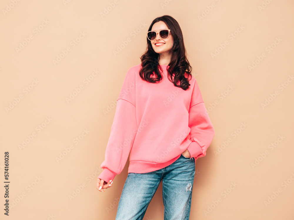Young beautiful smiling female in trendy summer jeans and pink sweater clothes. Sexy carefree woman posing near beige wall in studio. Positive model having fun. Cheerful and happy. In sunglasses Young beautiful smiling female in trendy summer jeans and pink sweater clothes. Sexy carefree woman posing near beige wall in studio. Positive model having fun. Cheerful and happy. In sunglasses