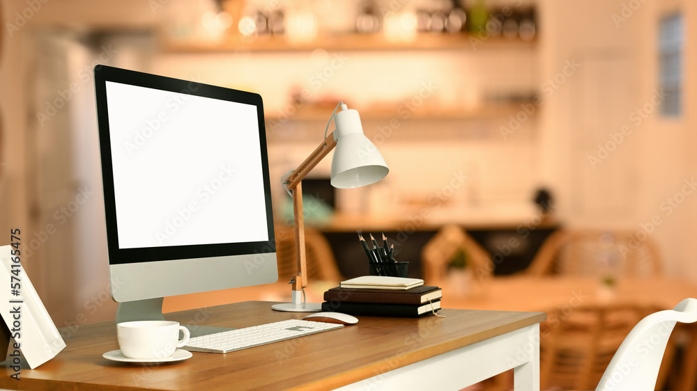 White blank screen computer monitor, lamp, coffee cup and books on table with blurred kitchen on background White blank screen computer monitor, lamp, coffee cup and books on table with blurred kitchen on background