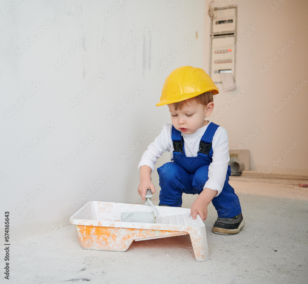 Adorable child construction worker crouching down and preparing paint roller for wall painting. Kid wearing safety helmet and work overalls while working on home renovation. Adorable child construction worker crouching down and preparing paint roller for wall painting. Kid wearing safety helmet and work overalls while working on home renovation.