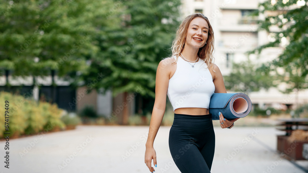 Joyful young caucasian girl holding yoga mat getting ready for workout standing outdoors Joyful young caucasian girl holding yoga mat getting ready for workout standing outdoors