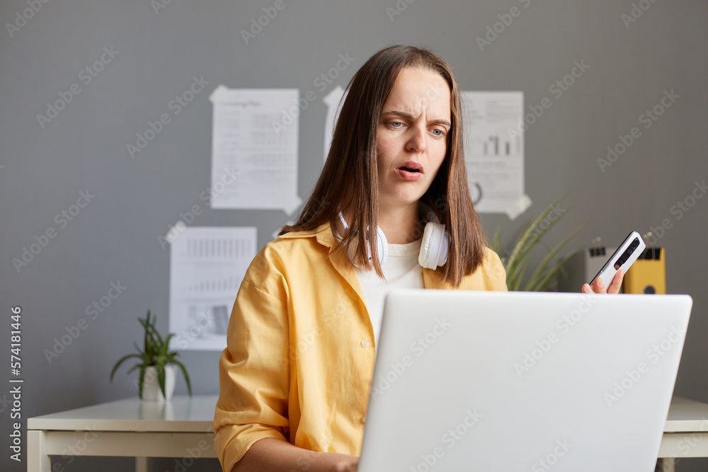 Image of shocked confused Caucasian woman with brown hair working on laptop, posing in office, holding cell phone in hands and looking at notebook display with big eyes. Image of shocked confused Caucasian woman with brown hair working on laptop, posing in office, holding cell phone in hands and looking at notebook display with big eyes.