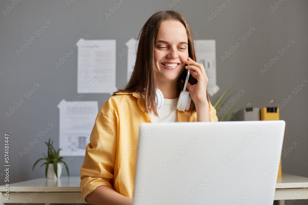 Pretty young adult woman working on laptop at her workplace in office, talking with client with charming smile, being pleased and friendly, trying to sell her service or works in call center. Pretty young adult woman working on laptop at her workplace in office, talking with client with charming smile, being pleased and friendly, trying to sell her service or works in call center.