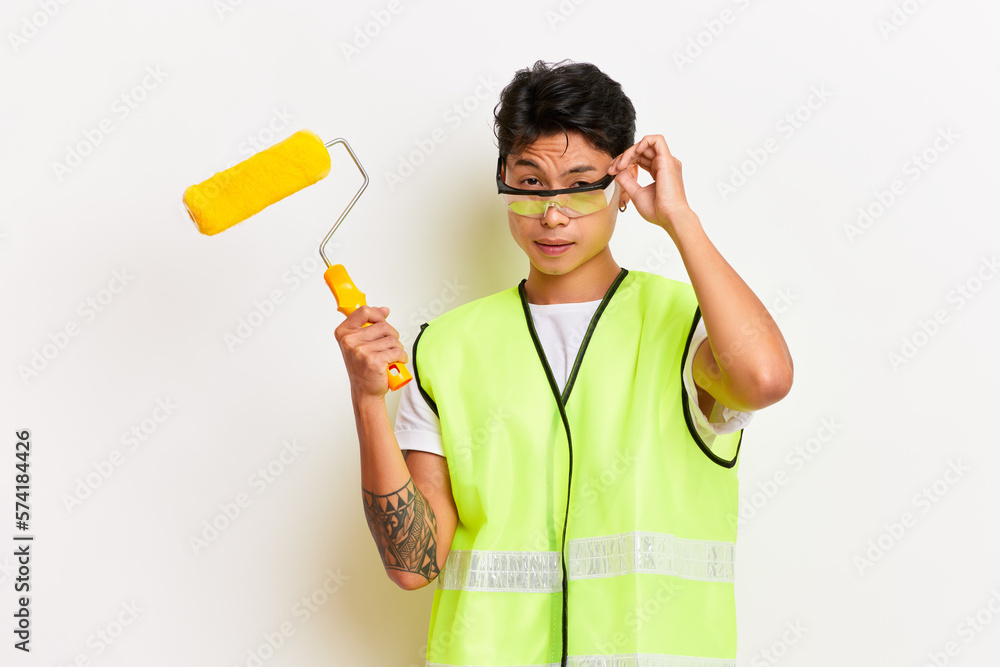 A shot of handyman holds paint roller wearing uniform and safety glasses isolated over white wall. People, manual work concept A shot of handyman holds paint roller wearing uniform and safety glasses isolated over white wall. People, manual work concept
