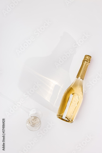 Top view white sparkling wine bottle and glass wine with sun shadow and glare on light white background. Flat lay  golden champagne bottle. Festive summer alcoholic drinks concept, trendy still life