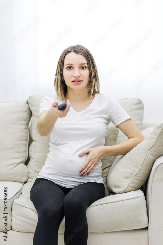 Pregnant woman holding a remote control sitting on the sofa.