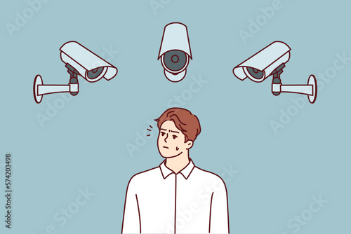 Embarrassed man stands among cameras recording every step of city dwellers to prevent commission of crimes. Dissatisfied man looking at CCTV cameras wondering if Big Brother is spying 