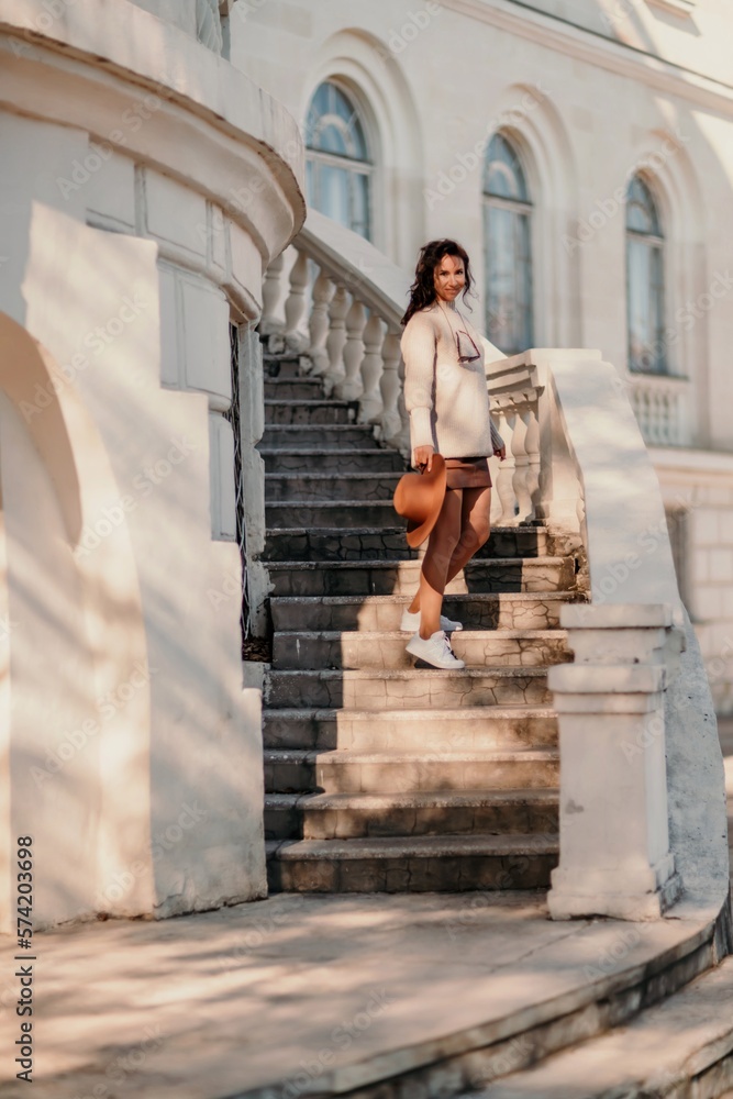 Woman walks in the city, lifestyle. Young beautiful woman in a loose light sweater, brown skirt and sneakers with a hat.