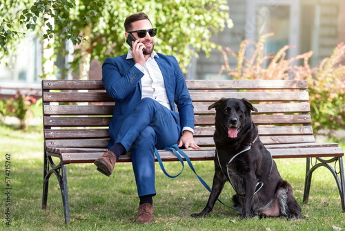 Handsome businessman in business suit is talking on his cell phone with a friend while walking his dog. Young man and his dog are sitting on bench in park. A break at work.