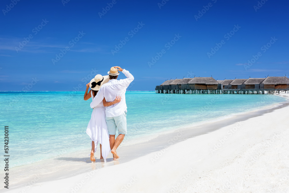 Happy couple in white clothing and with hats walks down a tropical beach with turquoise sea in the Maldives islands