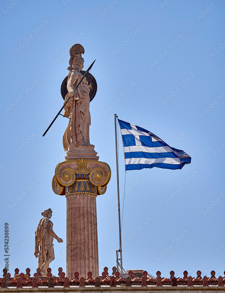 A view of the Greek flag, Apollo and Athena goddess statues represented as a warrior woman with helmet, shield and spear under clear blue sky. Travel to Athens, Greece.