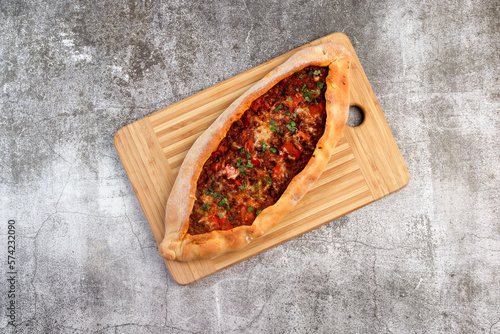 Pide (turkish pizza or pita dish) with ground beef, cheese, tomatoes and greens on a rectangular cutting wooden board on a dark grey background. Top view, flat lay