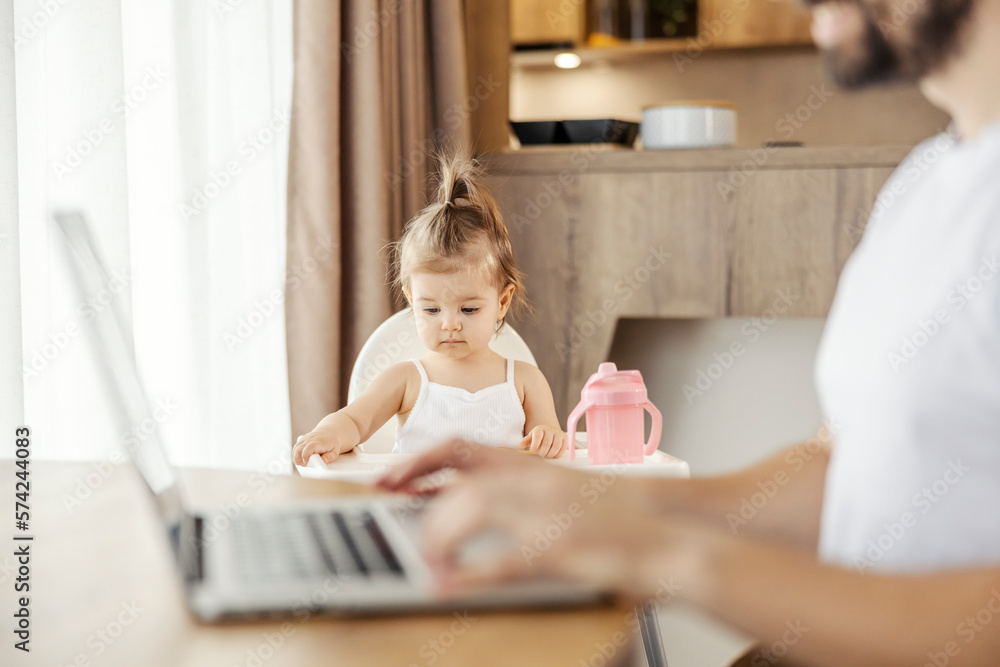A baby girl is sitting in her chair while her father is typing on a laptop in a blurry foreground. A baby girl is sitting in her chair while her father is typing on a laptop in a blurry foreground.
