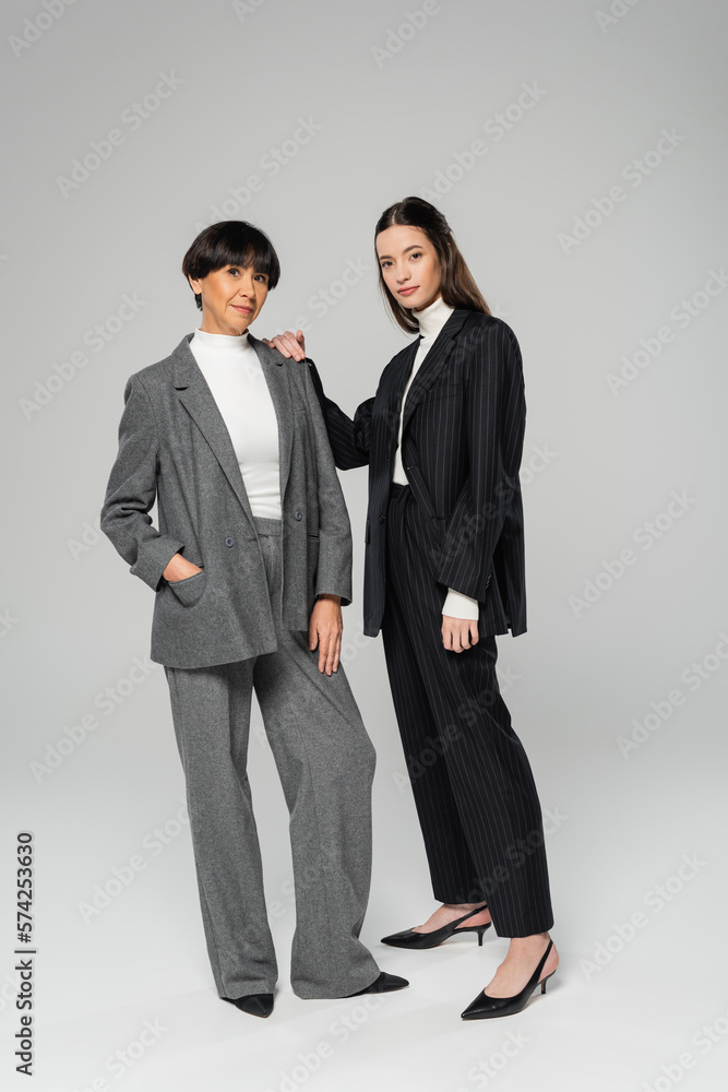 full length of asian mother and daughter in stylish pantsuits looking at camera while standing on grey background