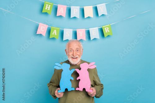 Portrait of attractive cheerful playful funky grey-haired man holding rabbits figure isolated over bright blue color background