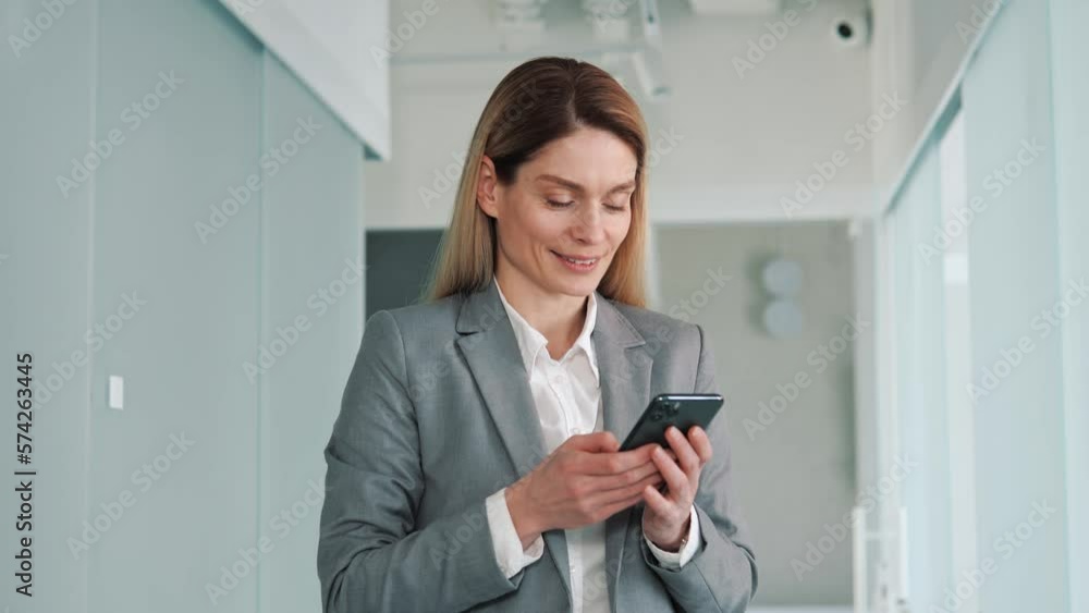 Happy confident young business woman standing in office hall looking into mobile phone smartphone screen chatting online in net smiling answering message in social media using gadget device, app. Happy confident young business woman standing in office hall looking into mobile phone smartphone screen chatting online in net smiling answering message in social media using gadget device, app.
