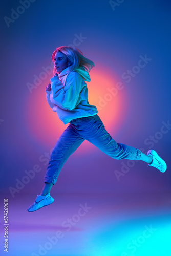 Active lifestyle, plans. Young woman in casual clothes, hoodie and jeans running over gradient studio background in blue neon light. Concept of emotions, facial expression, inspiration, sales, ad