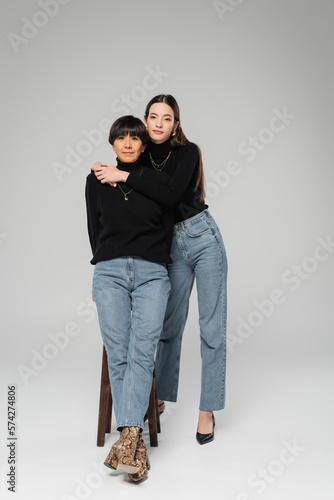 full length of fashionable woman embracing middle aged asian mother while posing on grey background