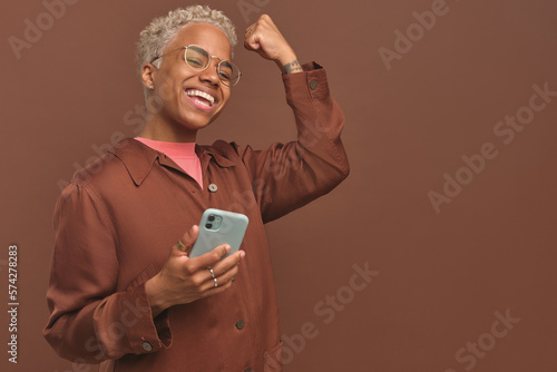 Young happy blonde African American woman makes gesture of victory after receiving SMS message in phone from bank with approval application for preferential loan or mortgage stands on brown background © antianti