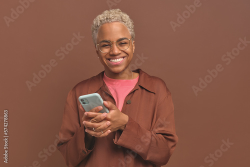 Young beautiful smiling African American woman holding phone with two hands and laughing at photo or video shared by friend on social network stands on brown studio background. Online communication