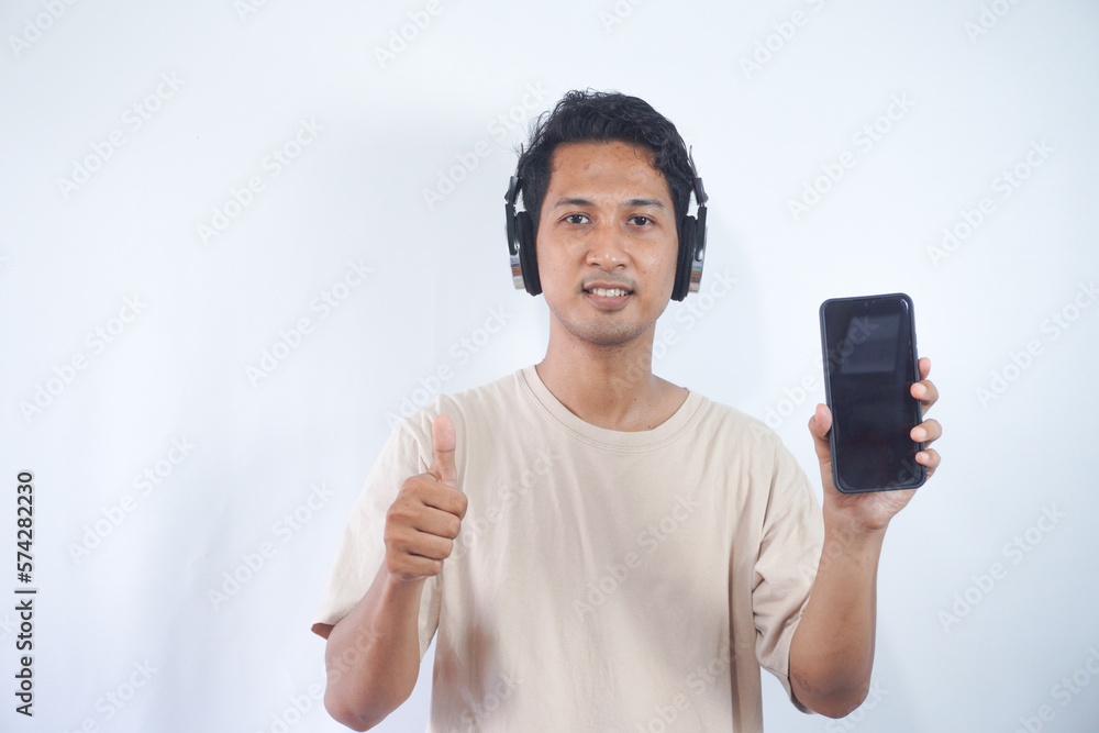 Half length shot of optimistic man holds smartphone with mock up screen, listens favourite soundtrack in headphones, wears casual cream t shirt, stands indoor. People and technology concept. Half length shot of optimistic man holds smartphone with mock up screen, listens favourite soundtrack in headphones, wears casual cream t shirt, stands indoor. People and technology concept.