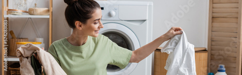 Smiling woman holding white and colorful clothes in laundry room, banner.