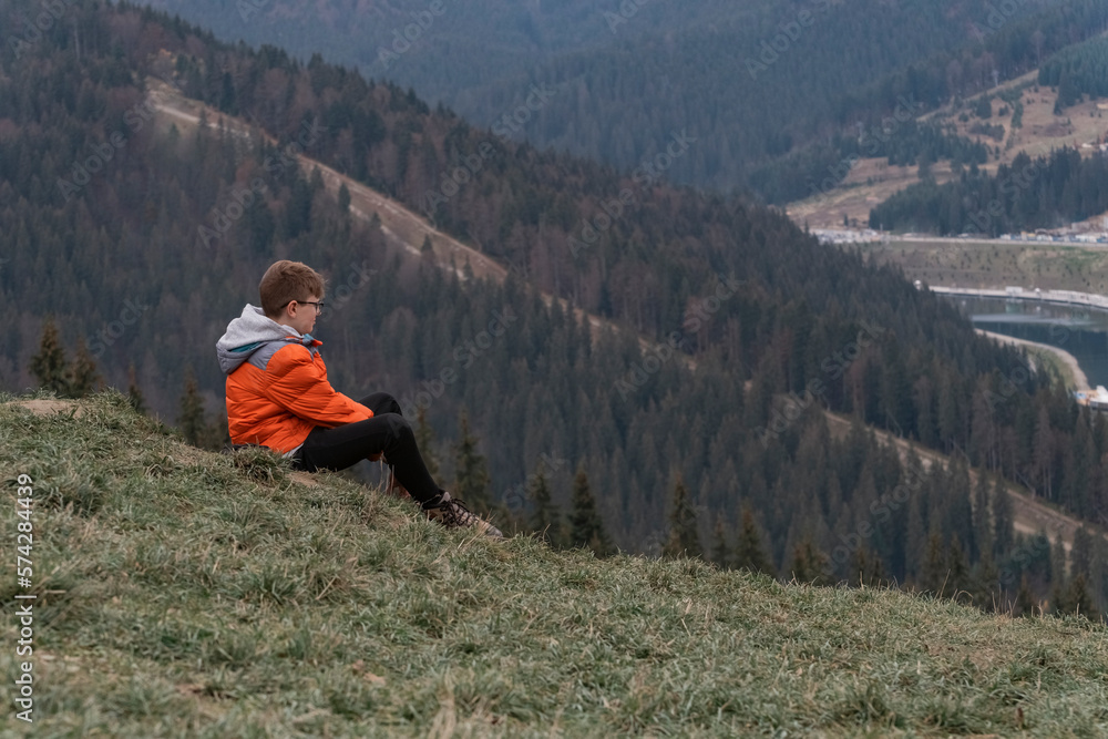 Teenage alone boy sits on hillside and looking at the distance, thinks about something. Travelling with children