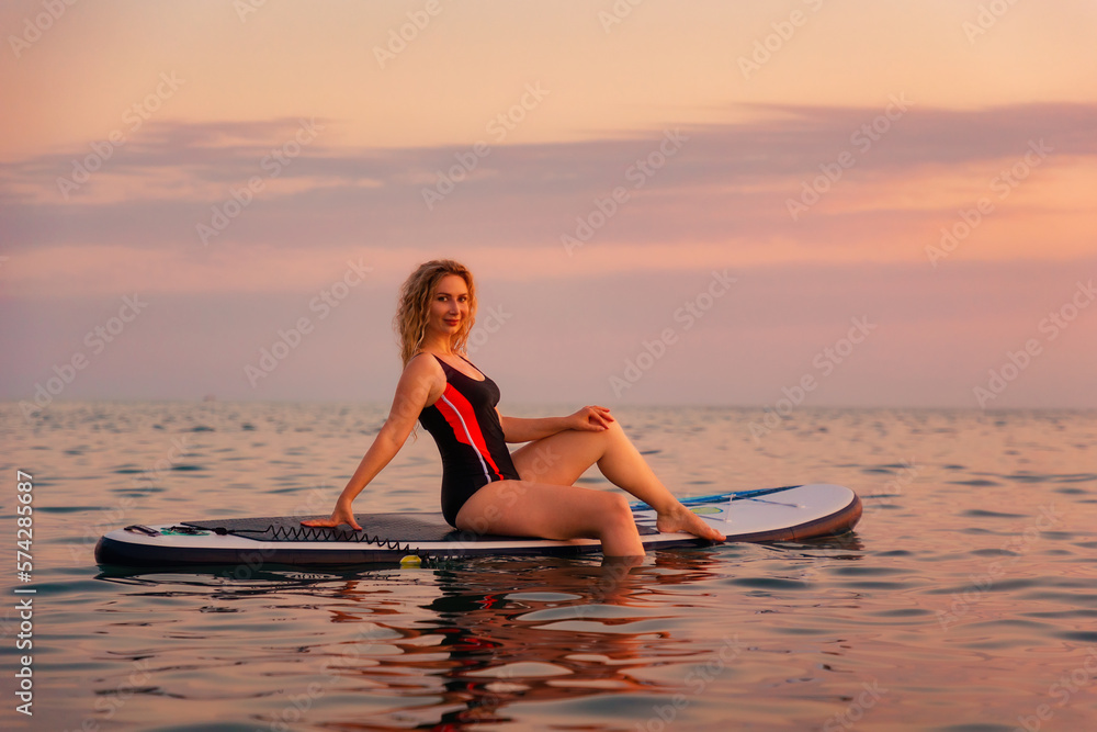 Summertime. Pretty young smiling woman posing sitting on sup board in the ocean. Sunset at the background. Copy space. The concept of sport and vacation