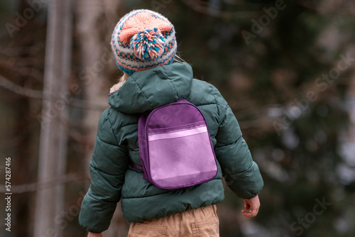 Little kid in knitted hat and jacket wears small backpack in pine forest. Child in fall in woods back view.