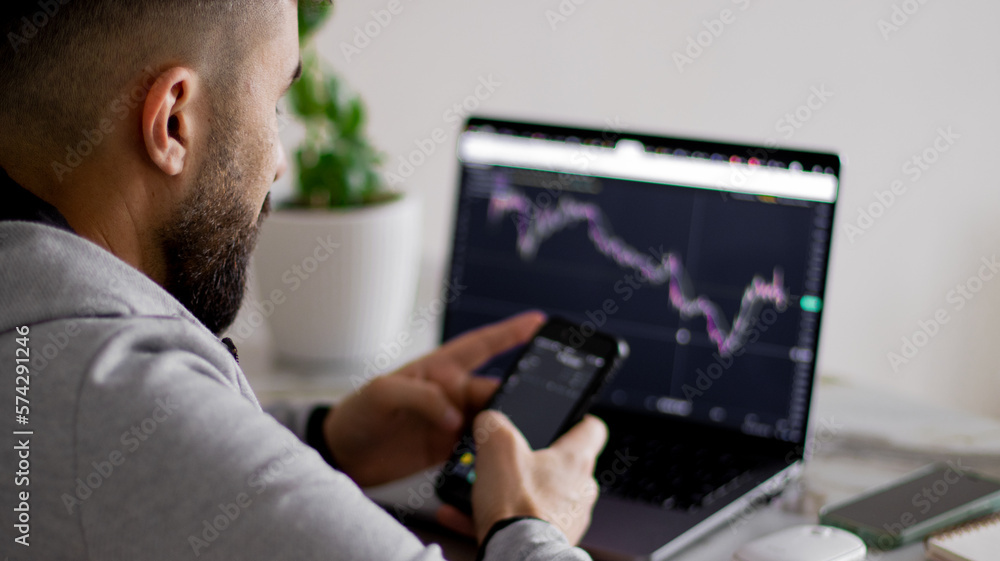 Happy bearded young man looks at the screen of his smartphone, sitting in front of laptop screen with graph of securities report. Businessman in casual clothes is working in the office on project. Happy bearded young man looks at the screen of his smartphone, sitting in front of laptop screen with graph of securities report. Businessman in casual clothes is working in the office on project.