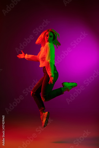 Young woman in casual clothes emotionally jumping, posing over magenta studio background in green neon light. Concept of emotions, facial expression, lifestyle, inspiration, sales, ad