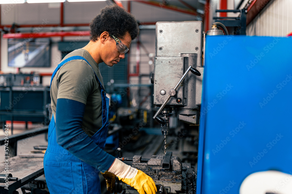A worker dressed in overalls and protective gloves, wearing safety glasses uses a machine to drill holes in a metal rod A worker dressed in overalls and protective gloves, wearing safety glasses uses a machine to drill holes in a metal rod