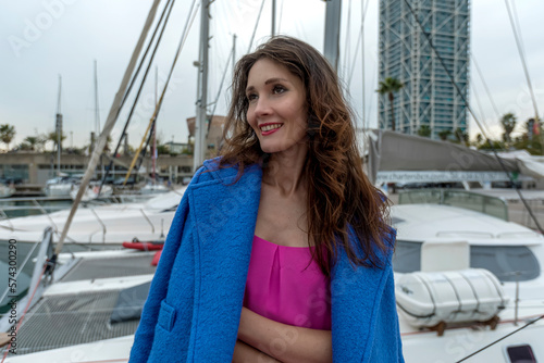 Beautiful woman in pink dress and blue jacket posing on a sailboat in a city harbor on a cloudy day.