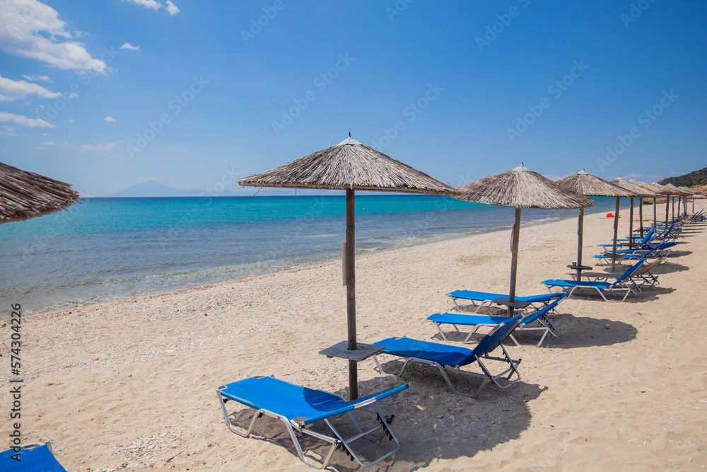 A tranquil beach scene with an idyllic horizon over the blue sea, a straw parasol for shade and a lounge chair to relax in - perfect for summer holidays.