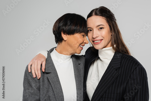 young woman in black blazer embracing happy asian mother while smiling at camera isolated on grey