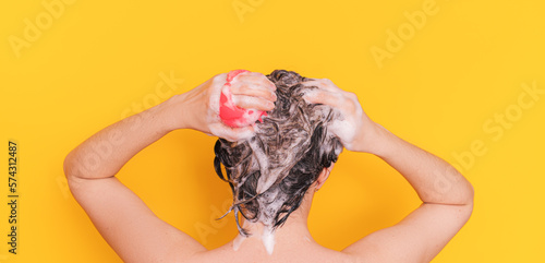 Lady with foam in her hair takes care of her scalp with pink color massager against yellow background.