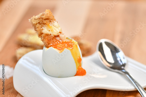 a single boiled egg in an egg cup, broken open showing yolk and with a slice of toast inserted with a spoon on a wooden surface