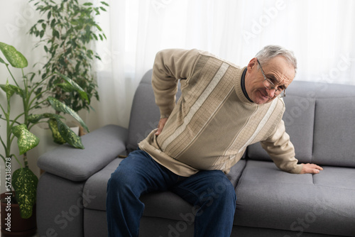 Caucasian old senior elderly unhealthy sickness grey male grandpa near sofa at home alone holding hands on back having emergency painful muscle and backache injury problem. © Angelov