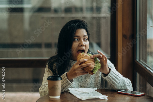 A young African American woman sits in a cafe and eats an appetizing burger. Bad cheap food. Wrong nutrition.