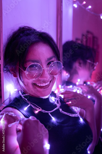 A young woman is smiling and posing next to the mirror wearing sunglasses surrounded by lights
