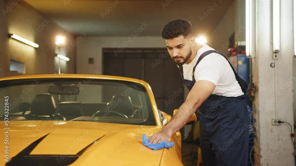 Portrait of car wash worker man, wearing protective clothes uniform overalls , cleaning modern yellow luxury car with microfiber cloth. Car detailing or vehicle concept. Cleaning modern luxury car. Portrait of car wash worker man, wearing protective clothes uniform overalls , cleaning modern yellow luxury car with microfiber cloth. Car detailing or vehicle concept. Cleaning modern luxury car.