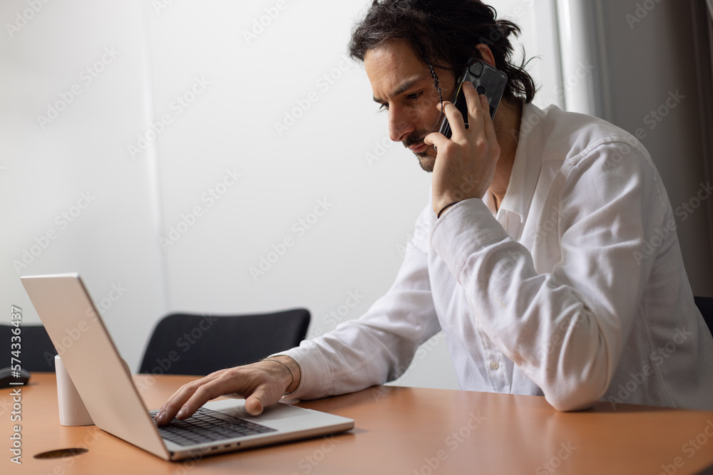 portrait d'un jeune homme souriant, employé de bureau assis devant son ordinateur au bureau. Il téléphone avec son smartphone portrait d'un jeune homme souriant, employé de bureau assis devant son ordinateur au bureau. Il téléphone avec son smartphone
