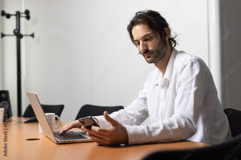 © Tof - Photographie - jeune homme souriant, employé de bureau assis devant son ordinateur au bureau. Il utilise son smartphone