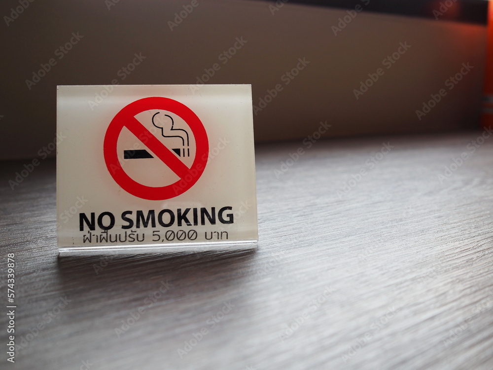 A white plastic sign placed on a wooden table inside the store reads NO SMOKING in black, with a picture of a cigarette in a red circle, and a text in Thai that reads, A white plastic sign placed on a wooden table inside the store reads NO SMOKING in black, with a picture of a cigarette in a red circle, and a text in Thai that reads,
