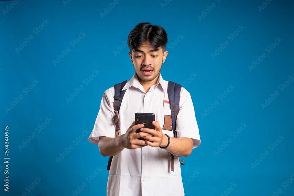 The expression of an Indonesian high school student who is surprised to see the smartphone he is holding against a blue background feels victorious, celebrating and winning The expression of an Indonesian high school student who is surprised to see the smartphone he is holding against a blue background feels victorious, celebrating and winning